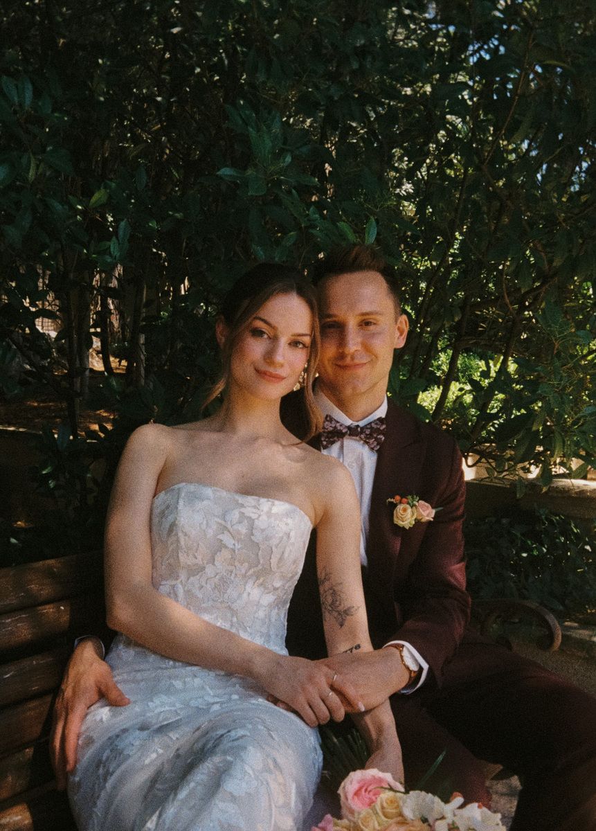 A bride in a strapless lace gown and groom in a burgundy suit pose together on a bench surrounded by lush greenery.
