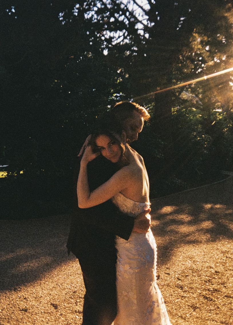 A couple embraces in golden sunset light, the bride in a lace gown and groom in a dark suit, surrounded by trees.