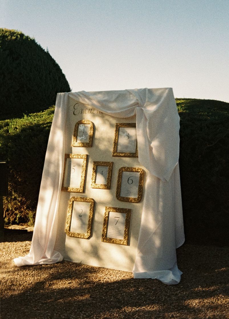 Elegant outdoor wedding seating chart display with gold ornate frames on white draped board, garden hedge backdrop.
