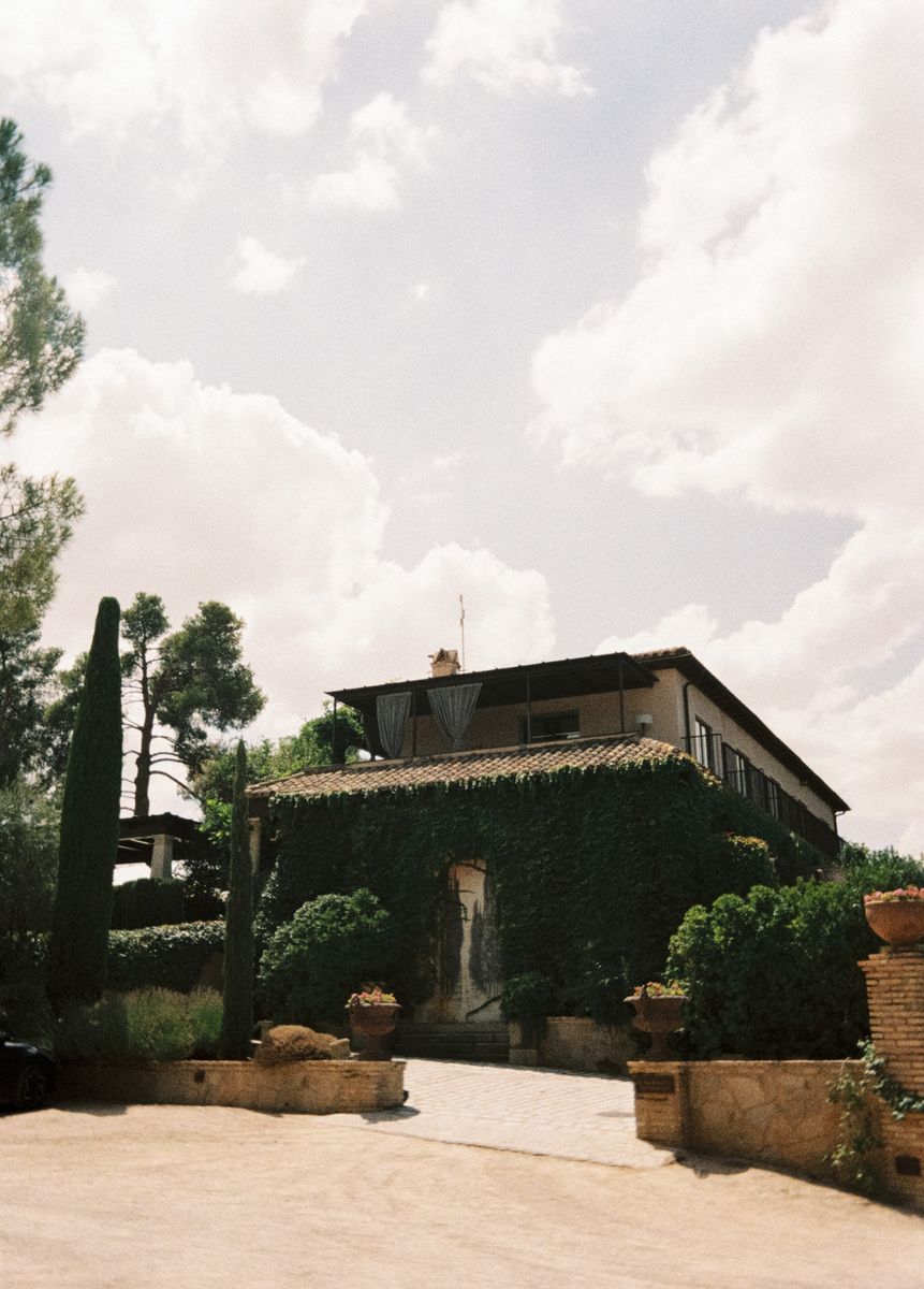 Ivy-covered Tuscan villa with terracotta roof, lush greenery, and cypress trees under a cloudy sky.