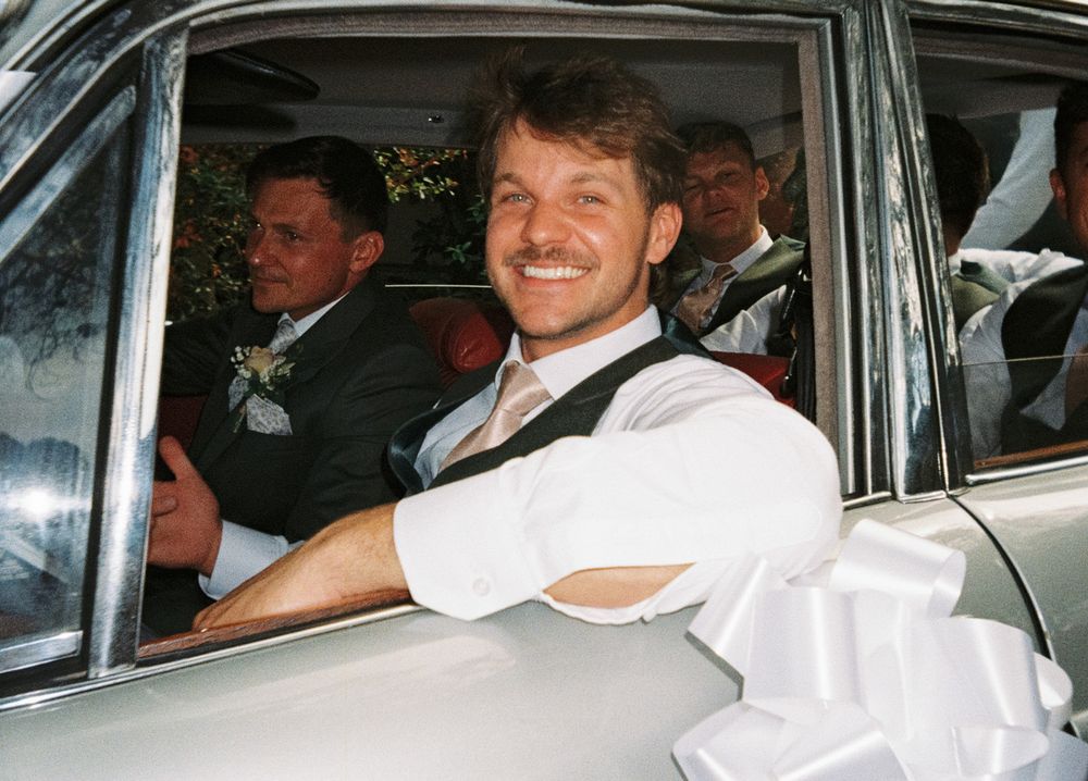 Smiling groom in white shirt and vest sitting in decorated wedding car with groomsmen in background.