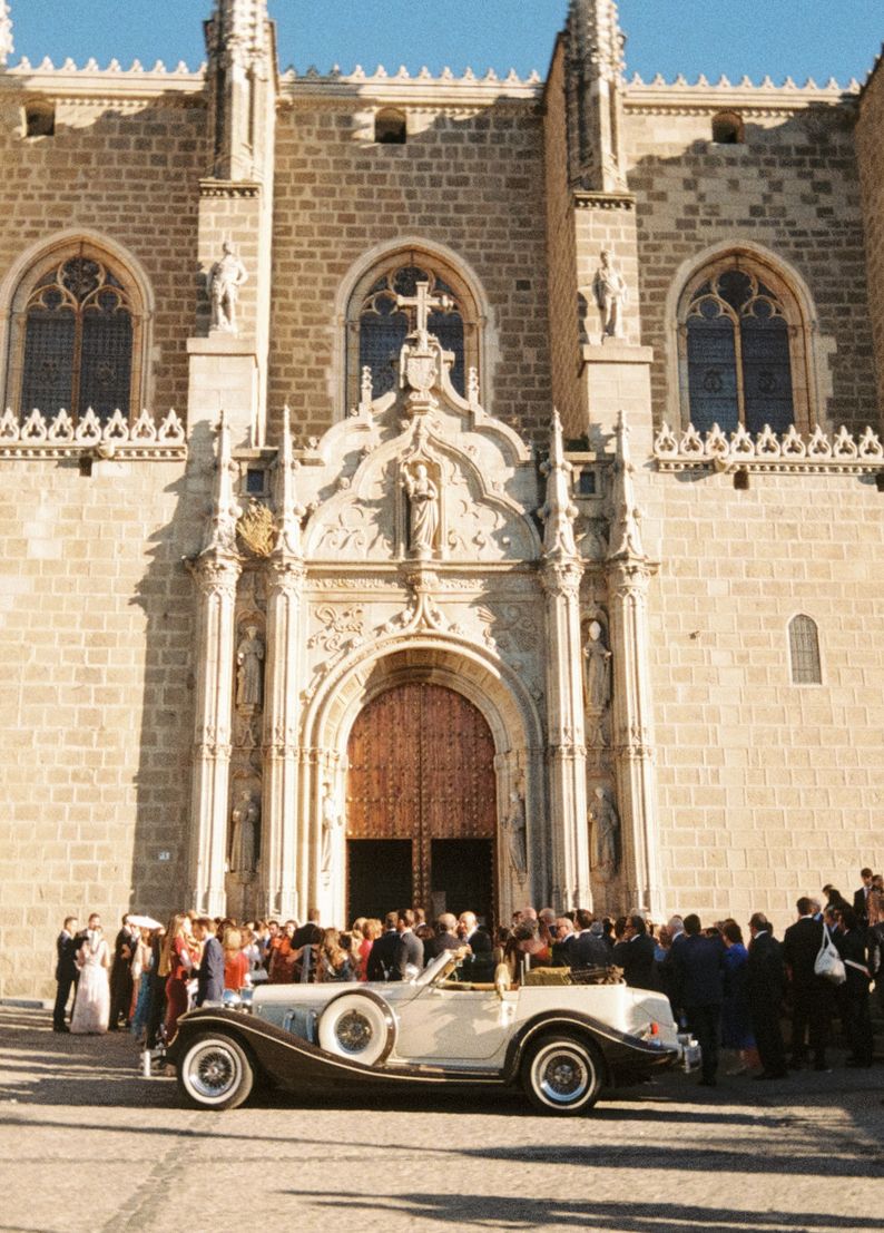 Vintage convertible car parked before ornate Gothic church entrance with wedding guests gathered outside.