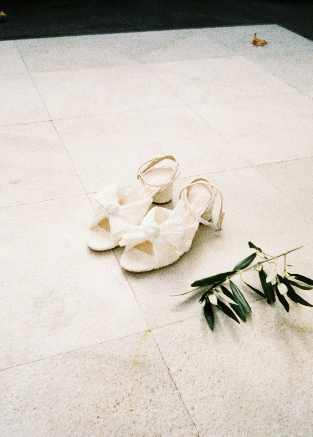 White bridal sandals with bow details sit on a light tile floor next to olive branches in an artistic composition.