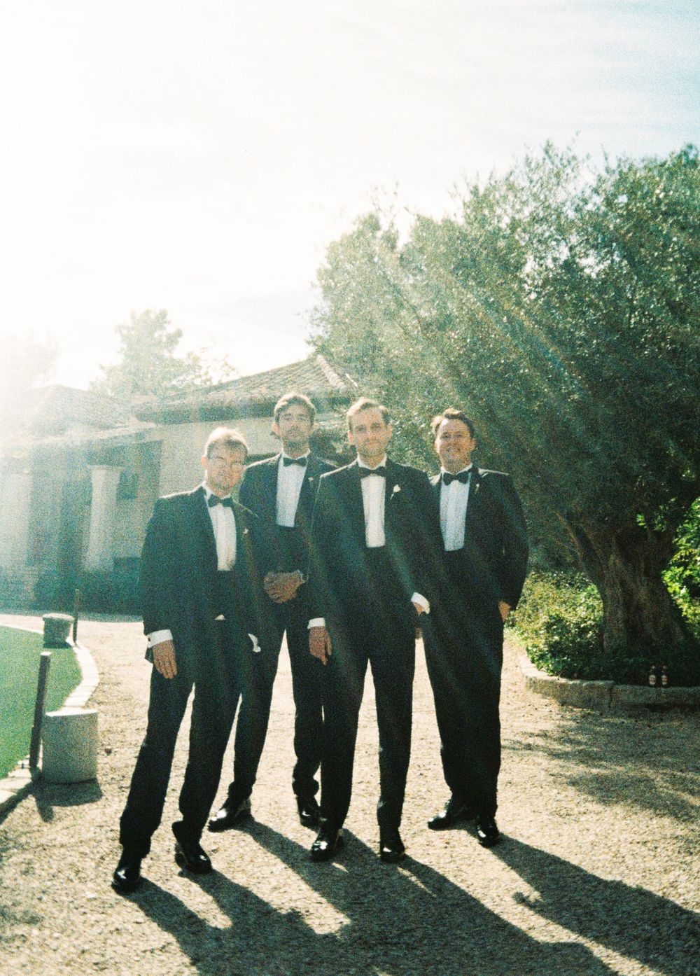 A group in black tuxedos stands together on a sunlit path with trees in the background.