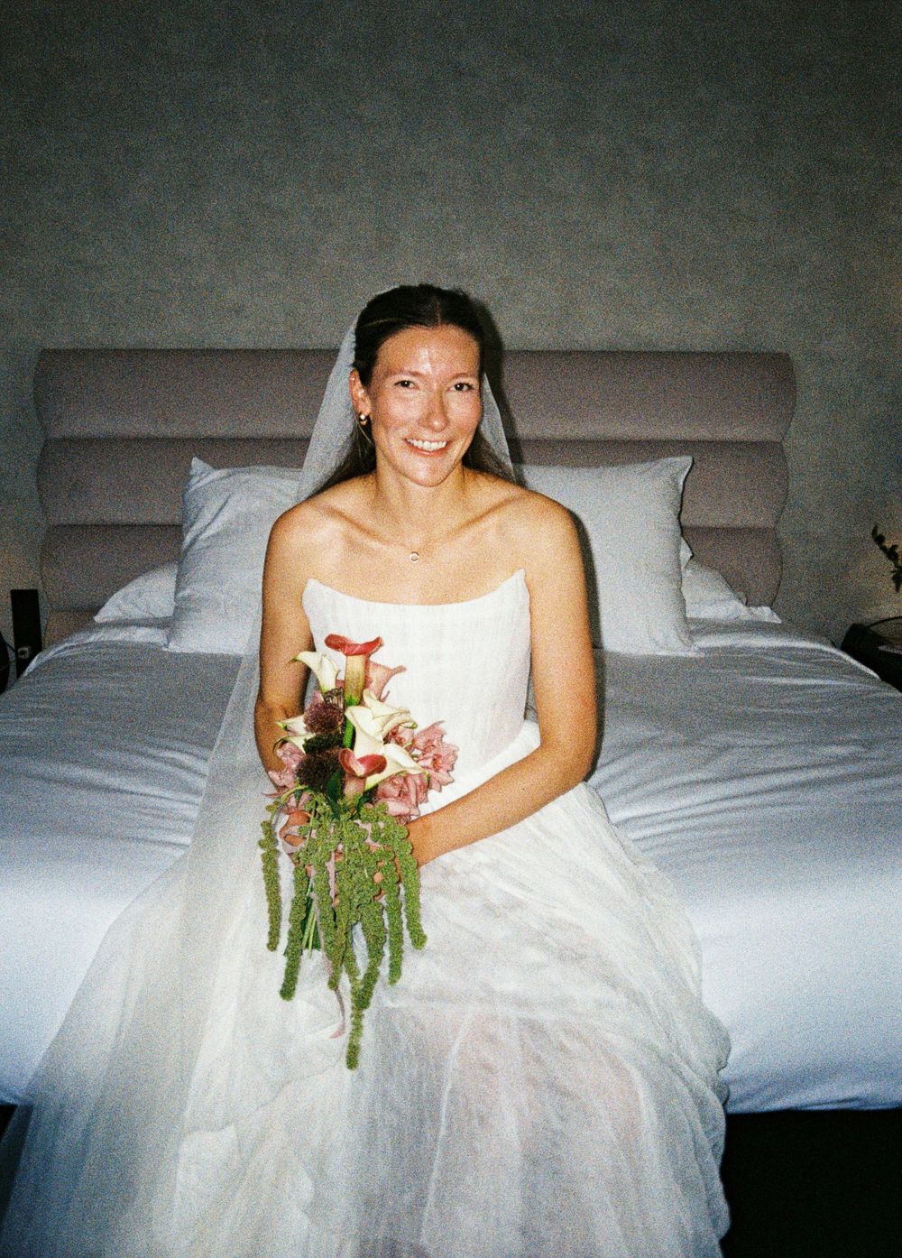A person in a white wedding dress sits on a bed holding a floral bouquet.
