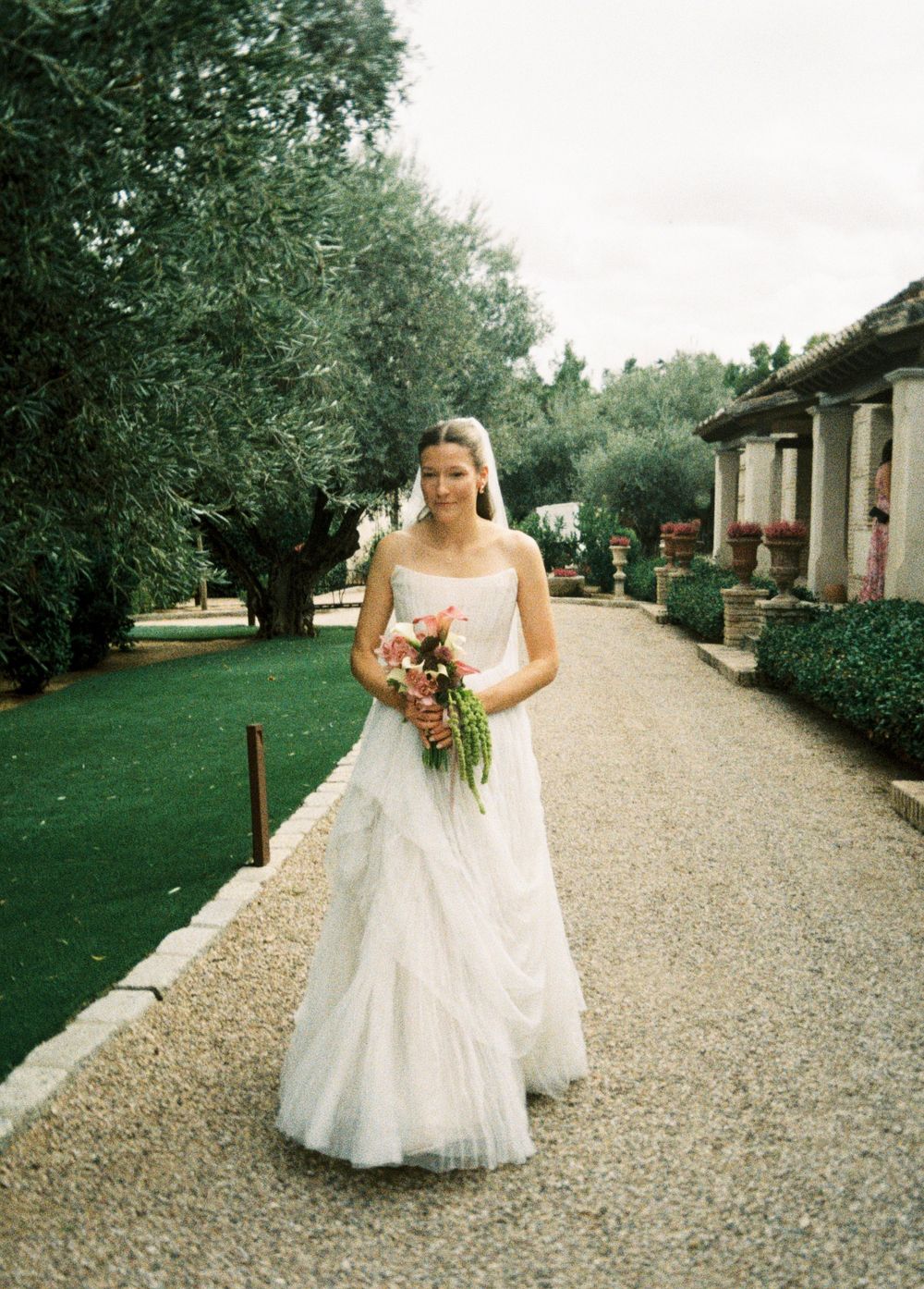 A bride in a flowing white gown walks along a gravel path lined with columns and lush greenery at an elegant venue.