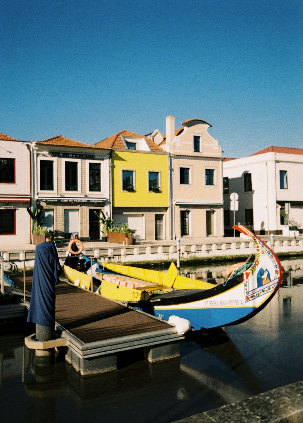 Colorful traditional boats line the waterfront of a Mediterranean village with white buildings and blue sky.