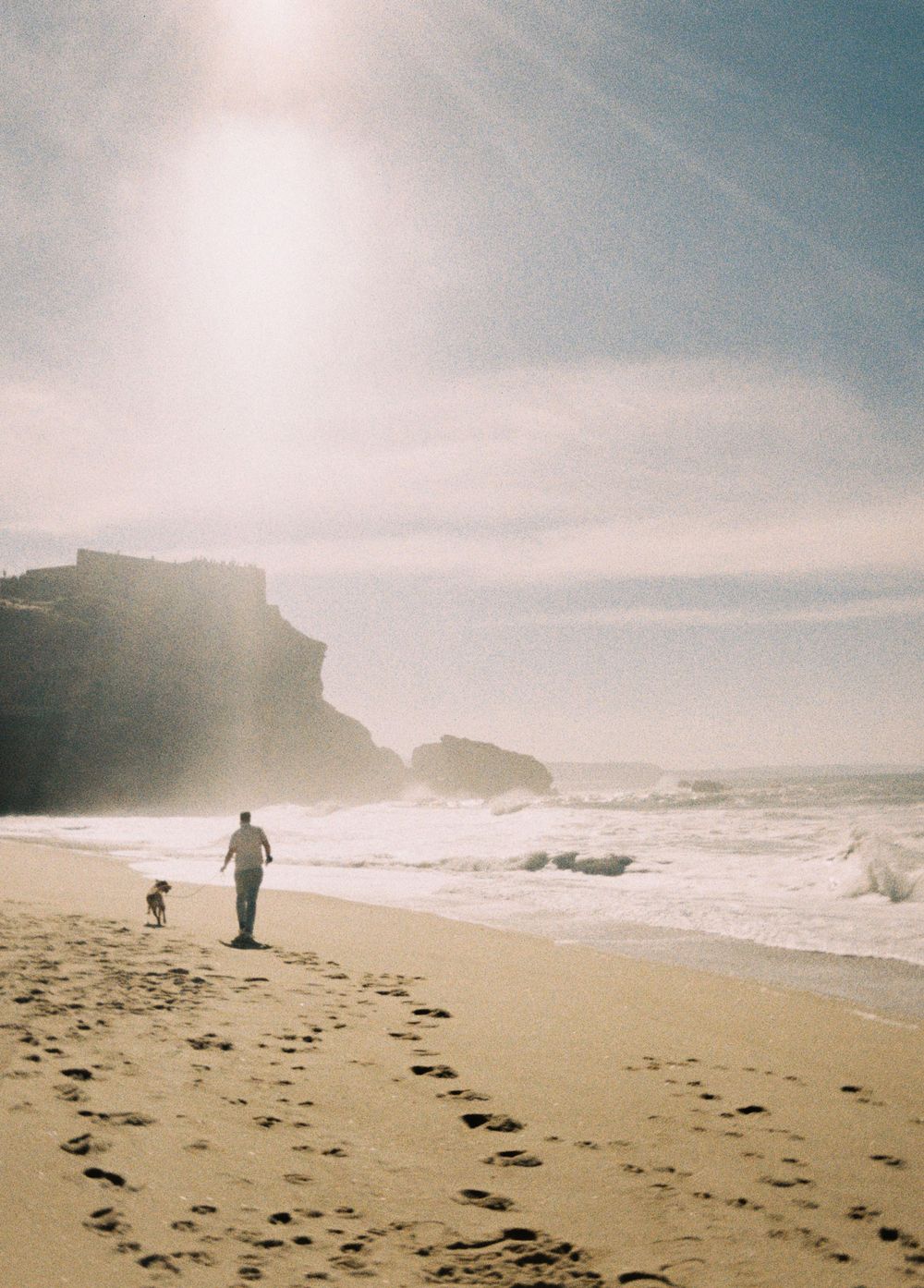 A figure and dog walk along a misty beach with cliffs and crashing waves in the background.