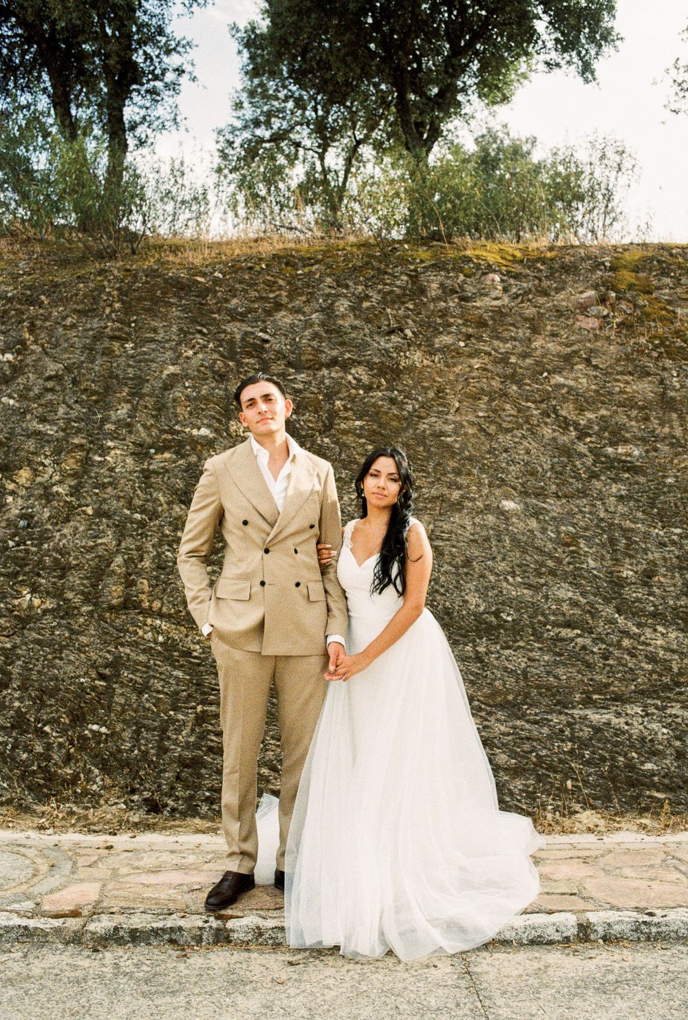 A couple in wedding attire poses against a rustic stone wall during an outdoor photo session.