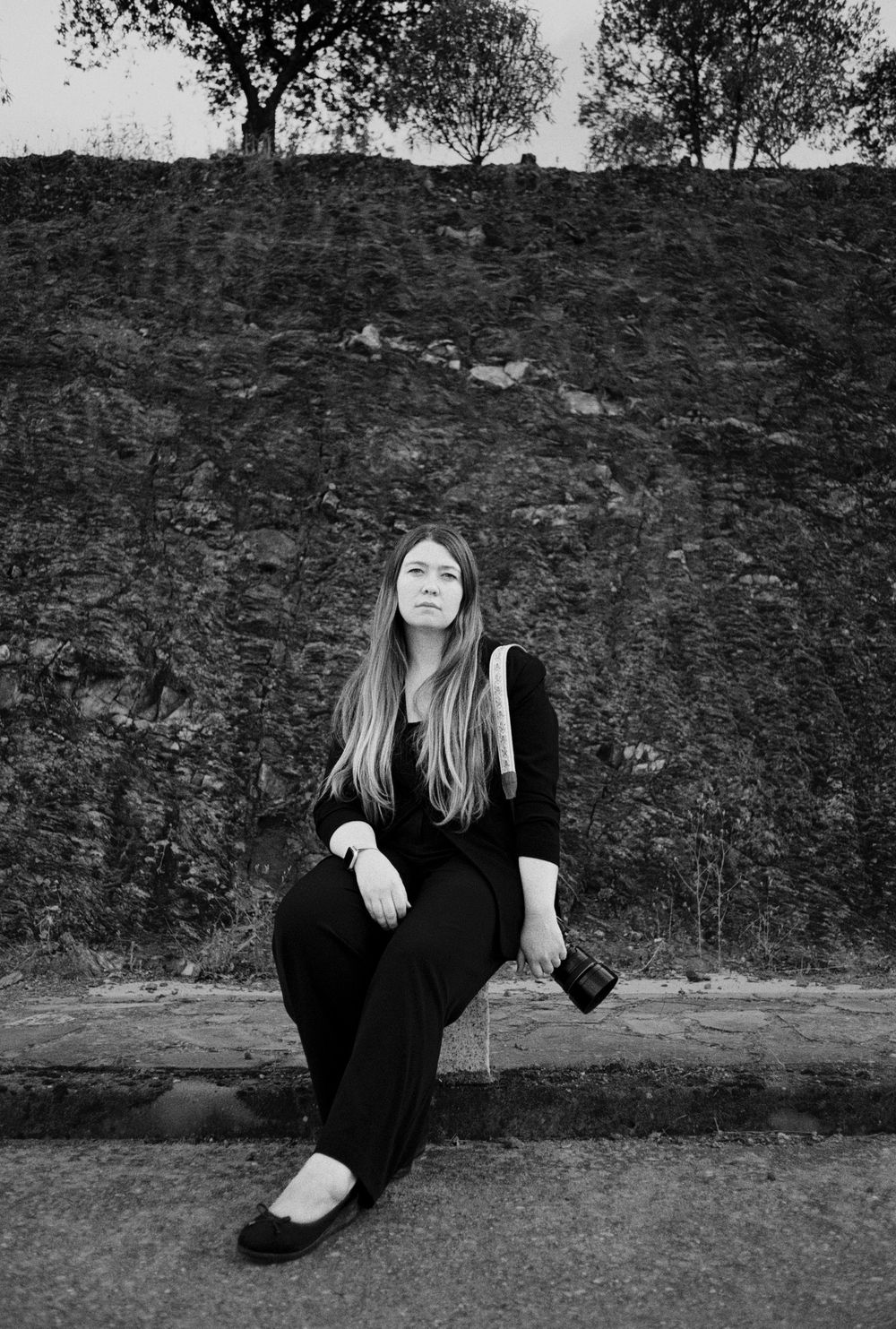 A black and white portrait of someone seated against a stone wall with trees visible above.