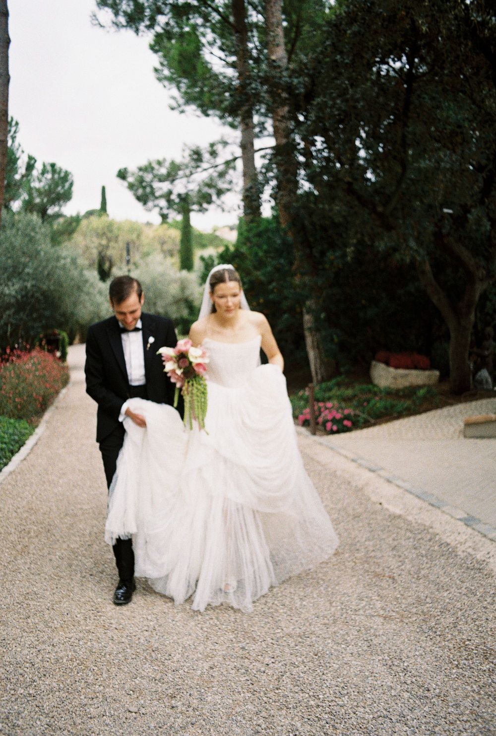 A couple walks along a gravel path surrounded by trees and flowers at an elegant outdoor wedding venue.