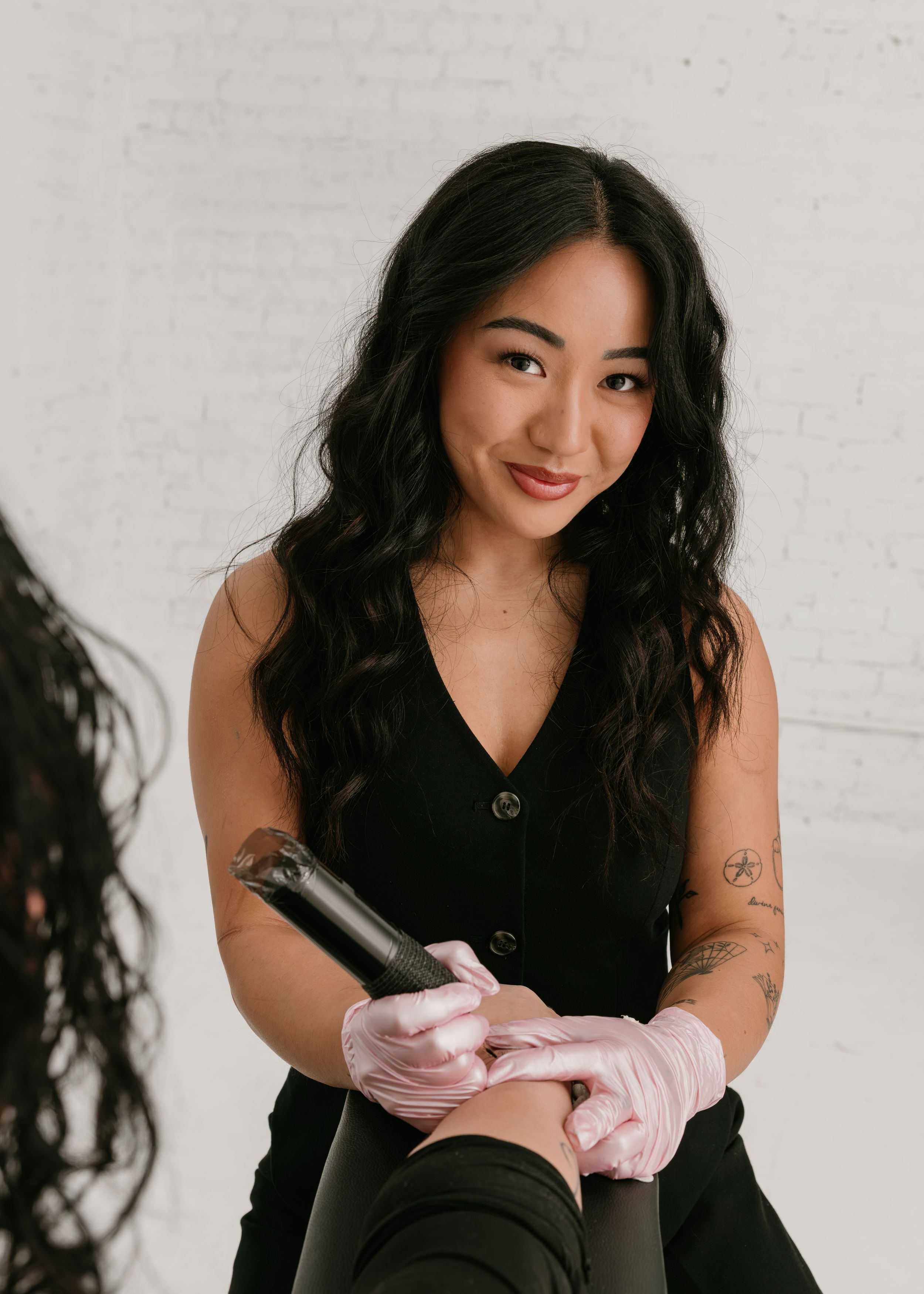 A person in black attire sits against a white wall while putting on pink protective gloves.