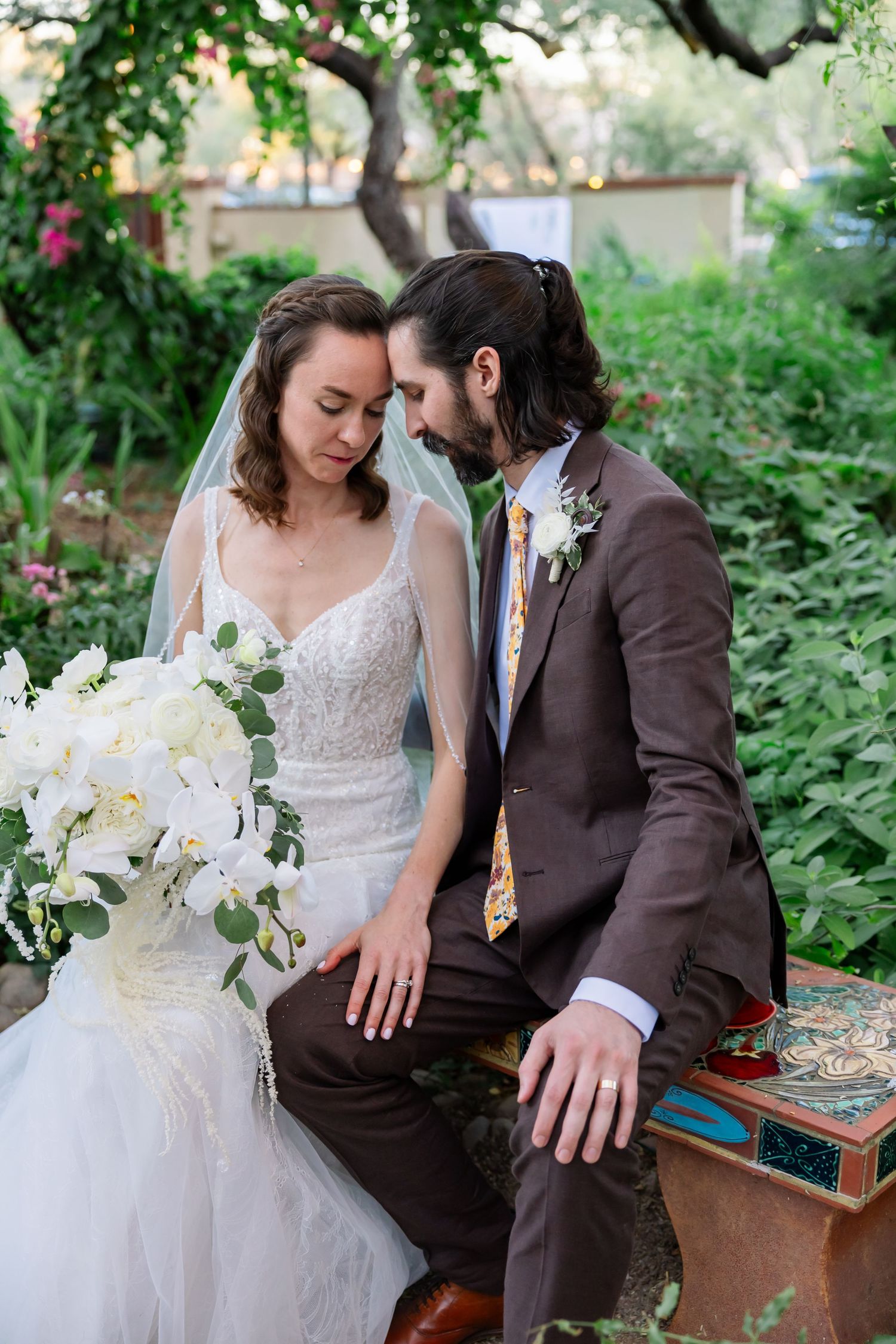 A romantic wedding couple shares an intimate moment on a brick wall patio surrounded by lush garden landscaping at sunset.