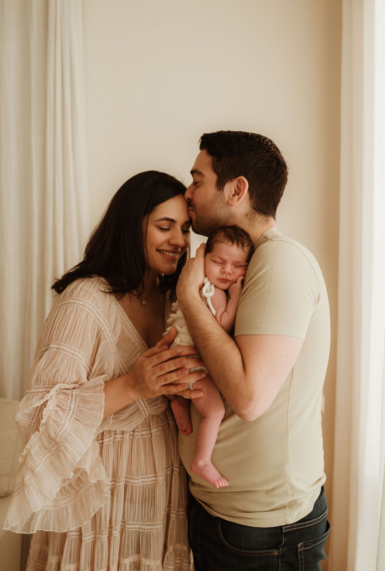 A small baby wrapped in a cozy white knit sweater lies on cream-colored fabric while a gentle hand rests on their head.