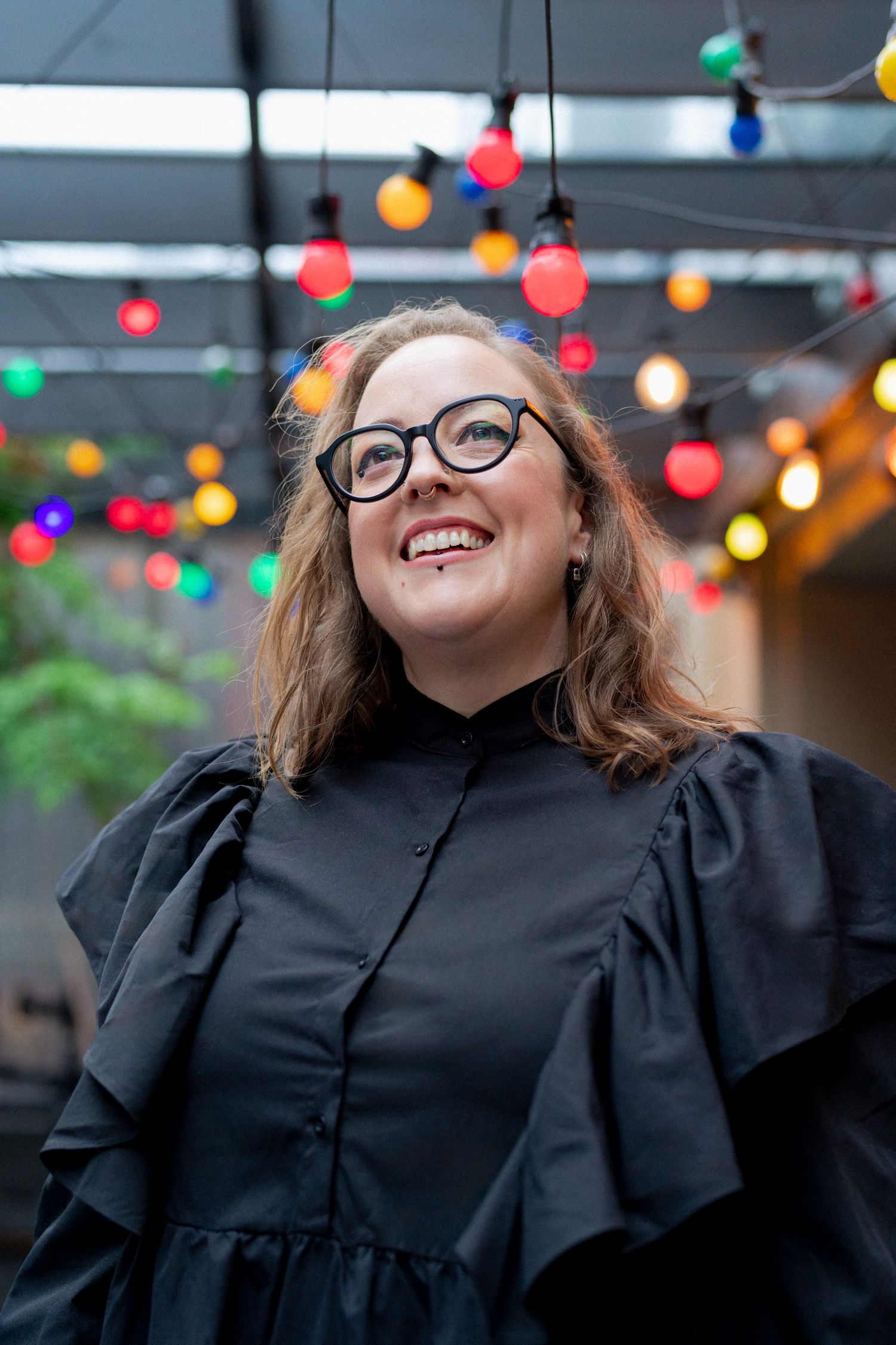 Emma Barnes has a beautiful big smile on her face, looking up at the bright festoon lights overhead. They wear a black dress & black glasses