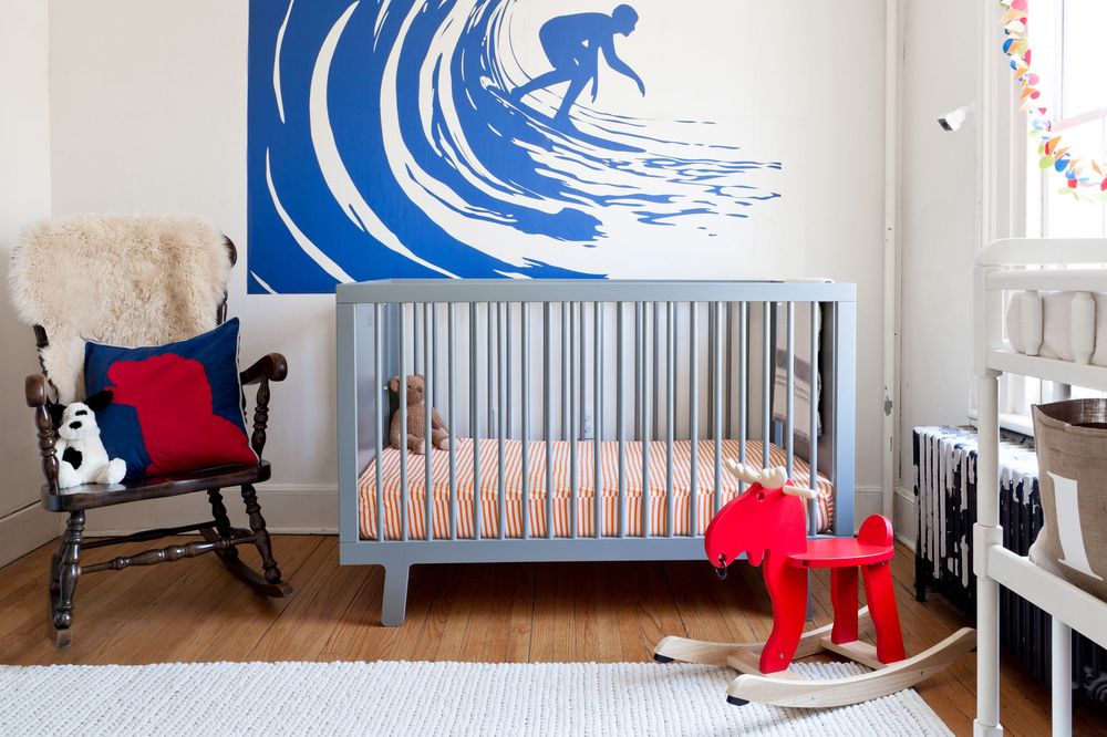 Modern nursery with blue surfing wall art, gray crib, rocking chair and red rocking horse decor.