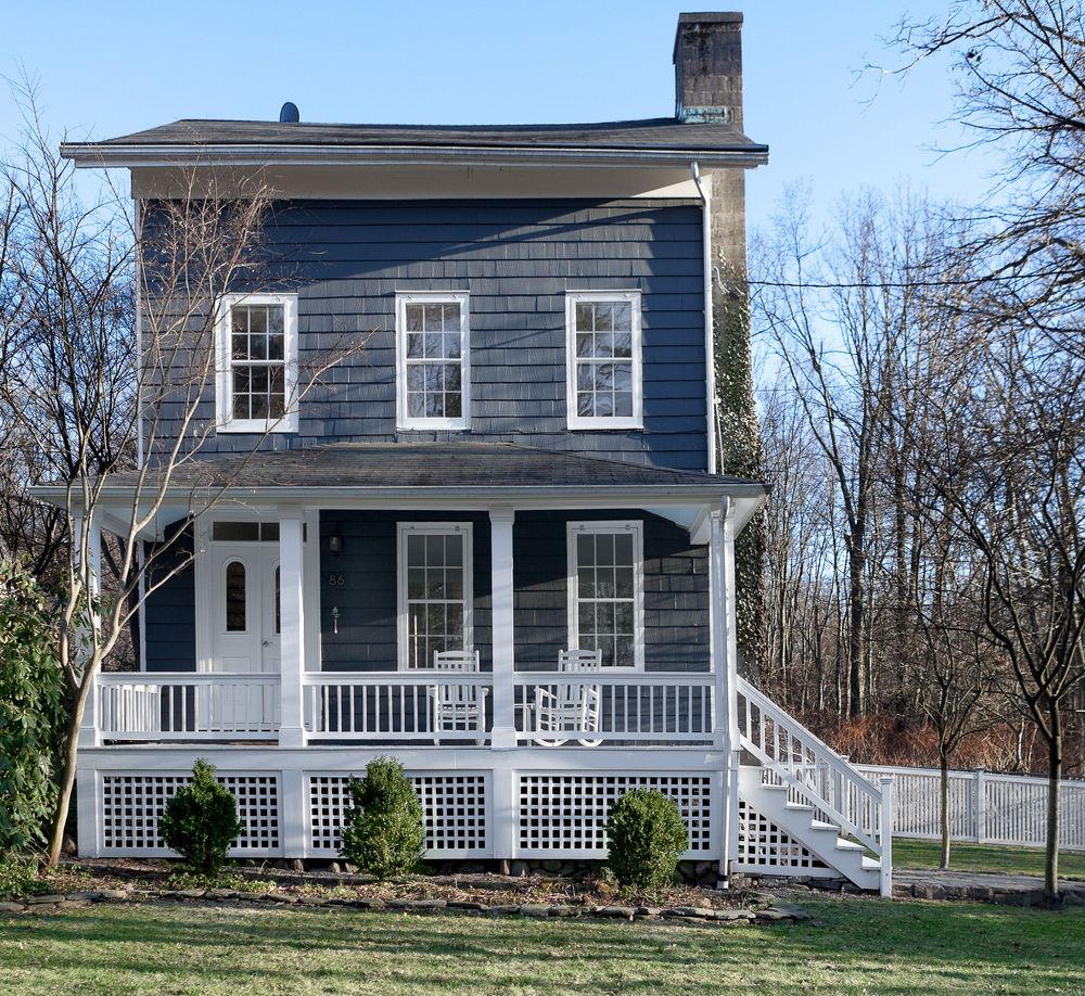 A classic white farmhouse with a wraparound porch and black shutters sits nestled among bare winter trees.