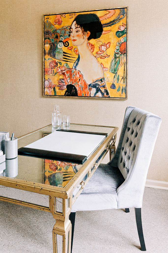 Elegant home office featuring a gold mirrored desk, tufted gray chair, and colorful Klimt-style artwork on beige wall.