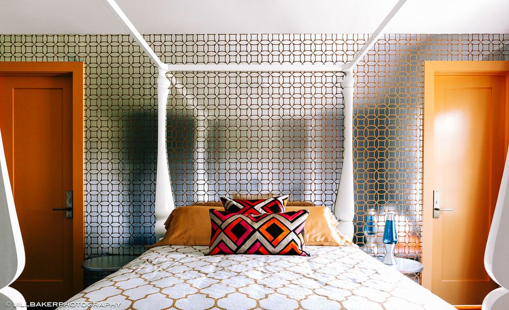 Modern hotel room interior with geometric patterned wallpaper and orange curtains illuminated by natural light.