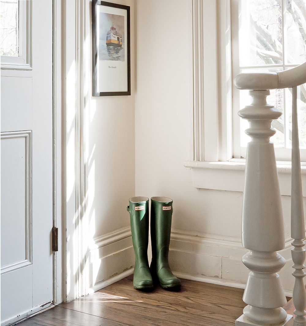 A pair of green rubber rain boots sits in a bright corner by a white door and window with elegant white molding.