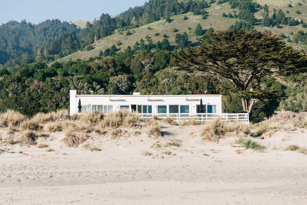 A panoramic view of a white modern beach house nestled against coastal hills along a sandy shoreline.