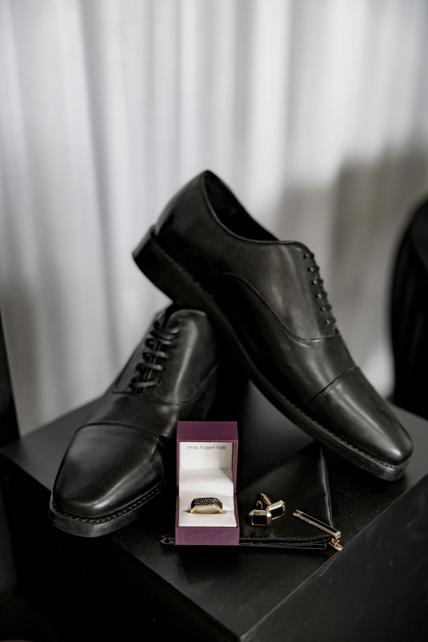 Black leather dress shoes and wedding rings displayed against white curtain backdrop.