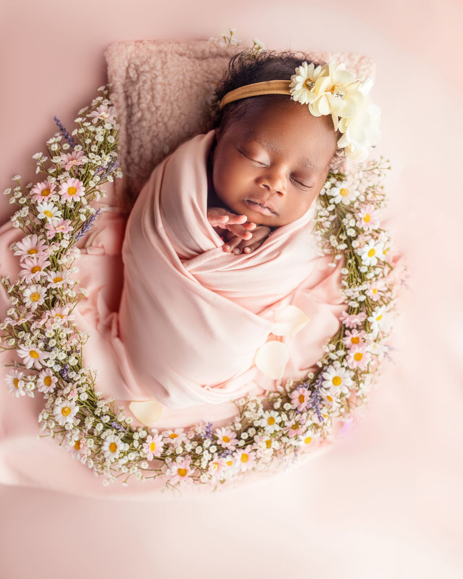 Sleeping newborn baby swaddled in pink wrap surrounded by delicate daisies and wildflowers.