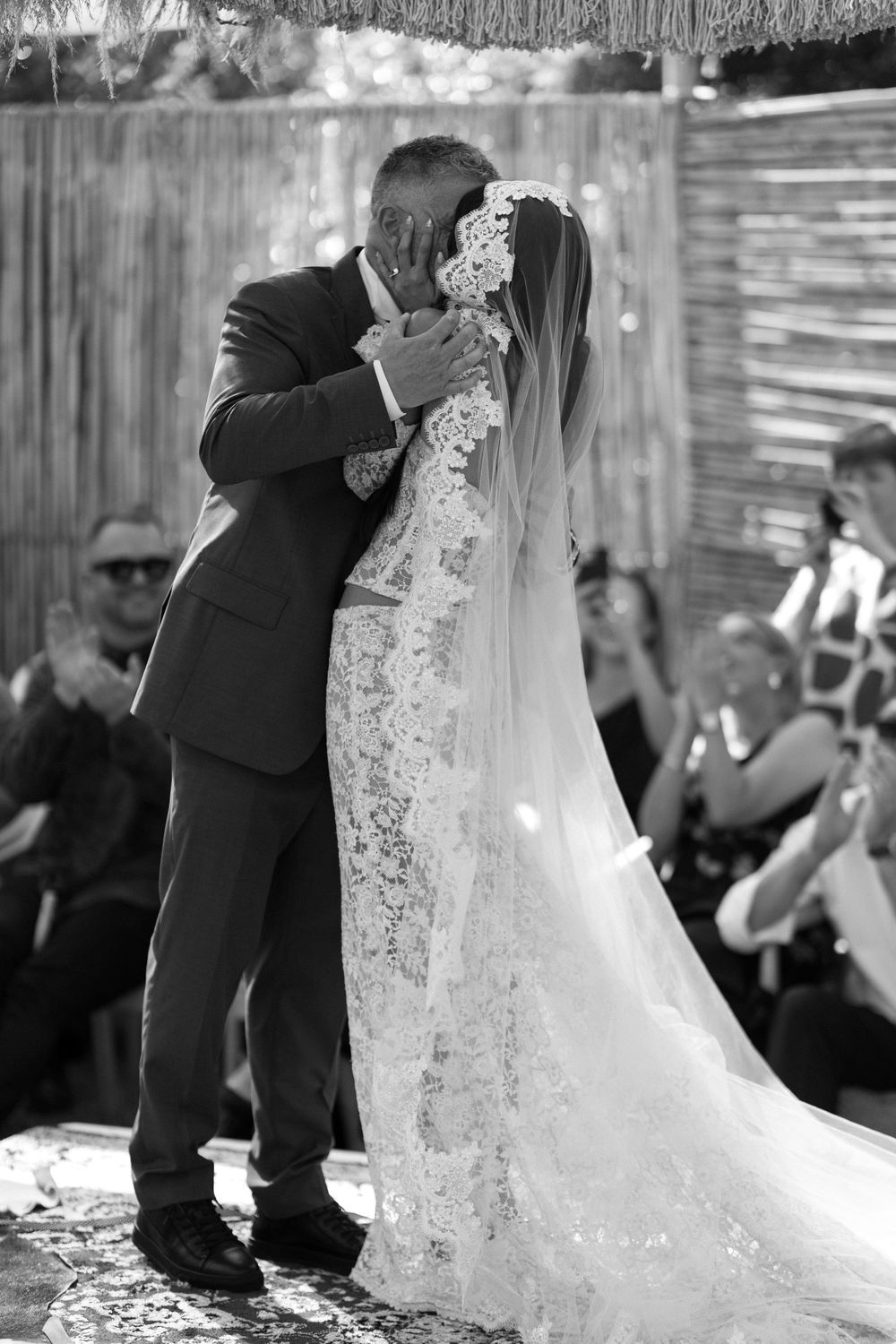 Wedding couple embracing during outdoor ceremony with lace dress and veil details.