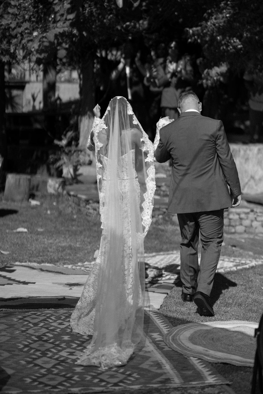 Black and white photo of newlyweds walking together with bride's lace veil trailing behind.