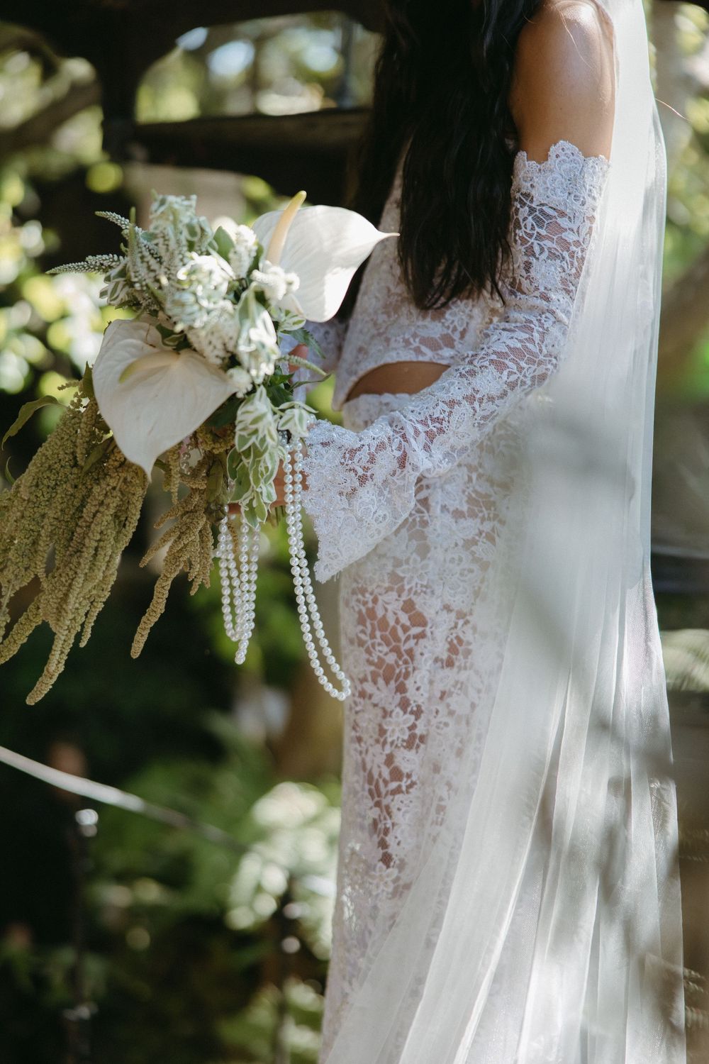 Elegant lace wedding dress with delicate pearl details and cascading white floral bouquet in natural lighting.