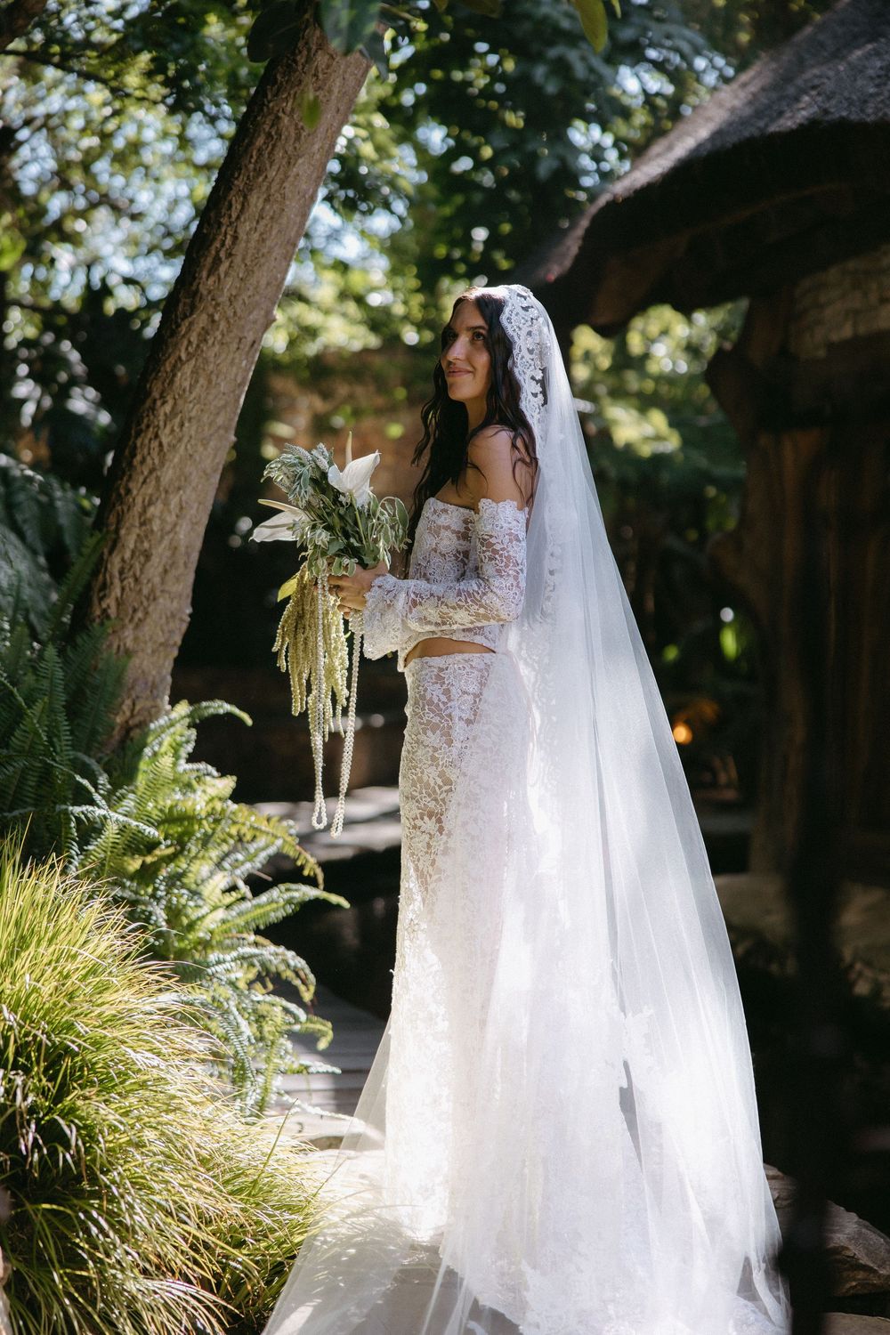 Bride in off-shoulder lace gown holds bouquet in garden setting with flowing veil.