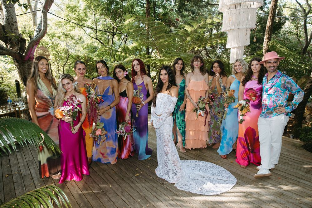 Large group of wedding guests in colorful dresses pose together on an outdoor deck surrounded by greenery.
