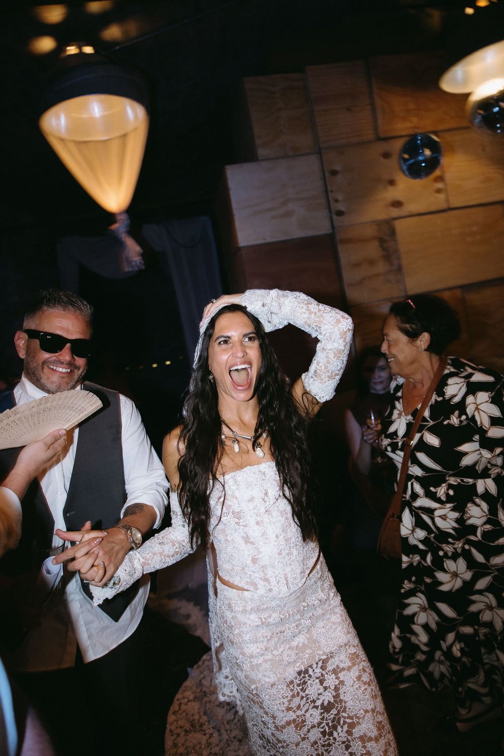 Wedding party dancing and celebrating with bride in lace dress under dim lighting.