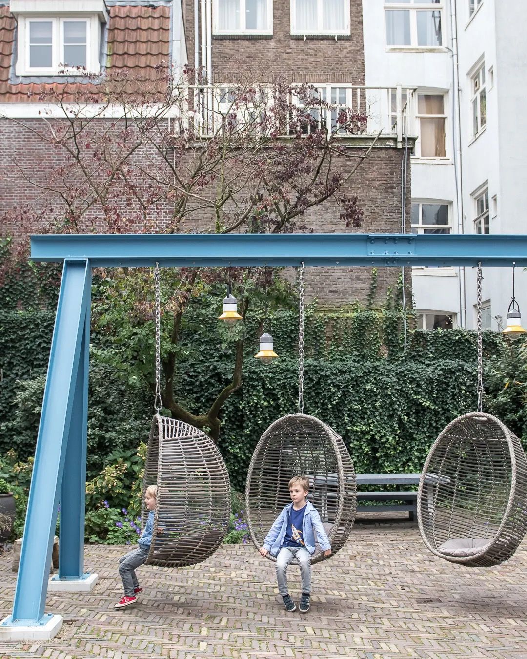 Children sitting on a basket swing in a small neighbourhood playground with city homes behind.
