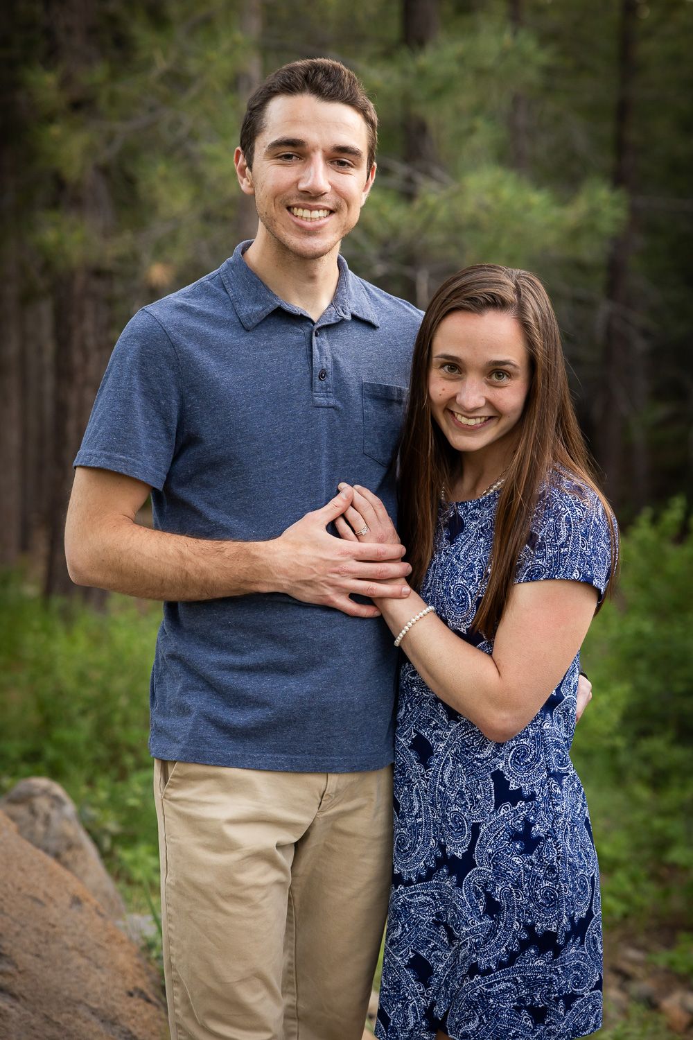 A couple in coordinated blue and khaki outfits poses together outdoors in a wooded setting for family photos.
