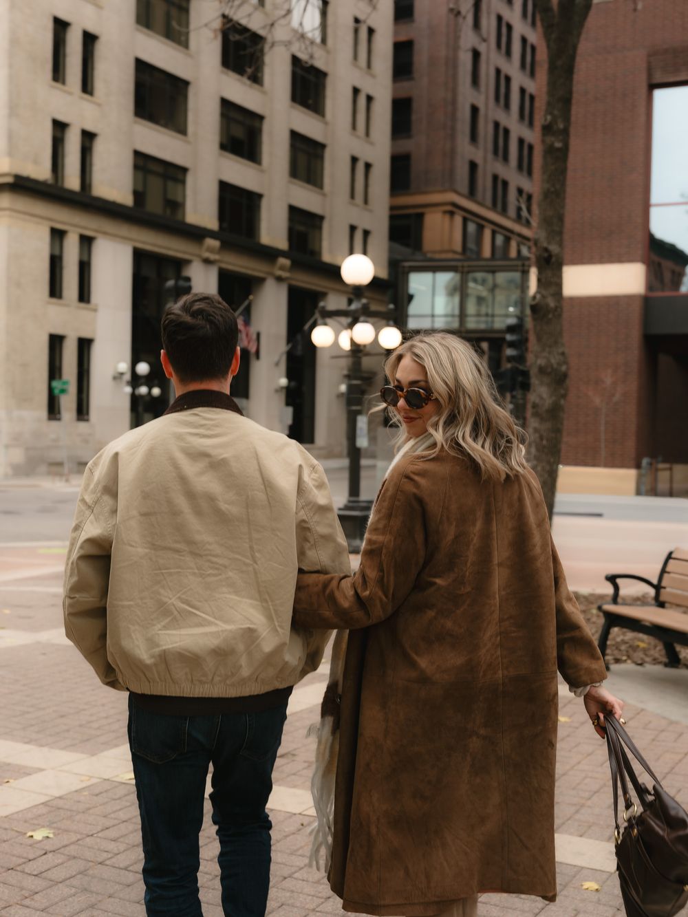Two people in casual attire walk together along a city sidewalk with tall buildings visible in the background.