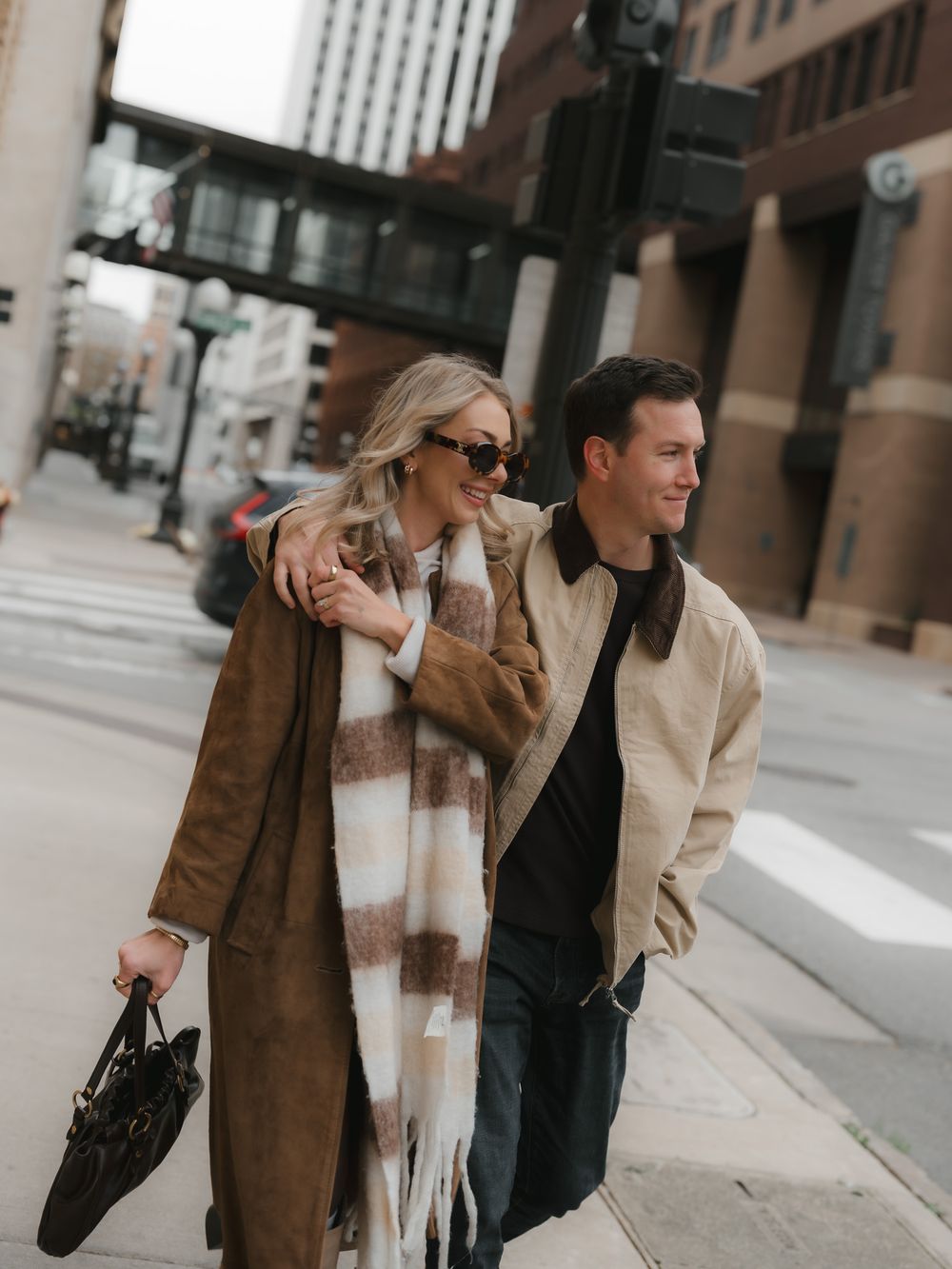 Two people stroll along a brick building exterior in coordinated beige and brown outerwear.
