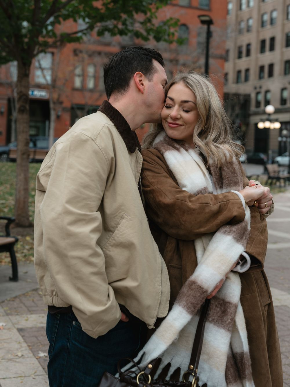 A romantic couple embraces on a city sidewalk, wearing warm coats on an autumn day in front of brick buildings.