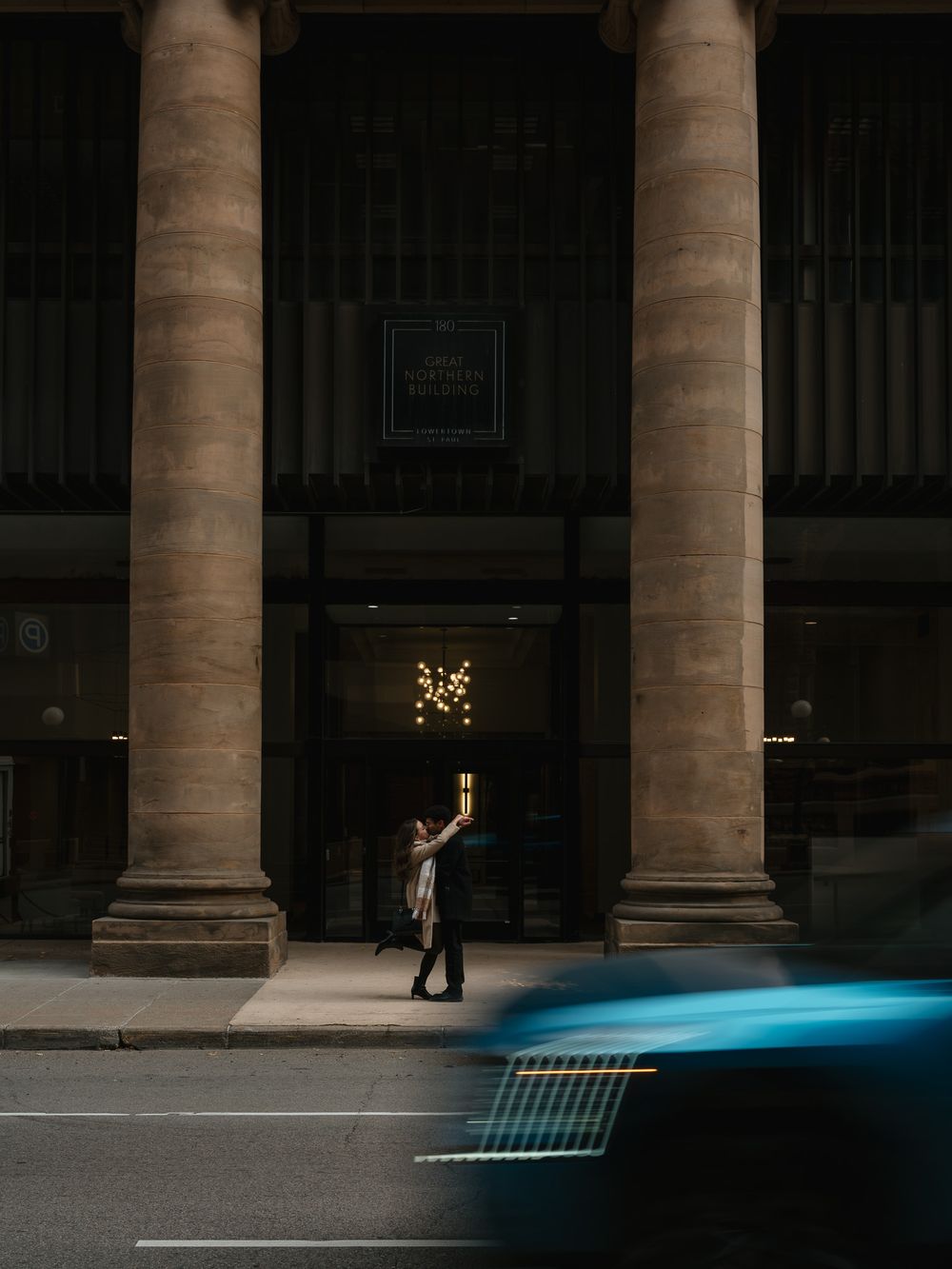 A silhouetted figure stands between grand columns as a blurred blue car passes by on the street.