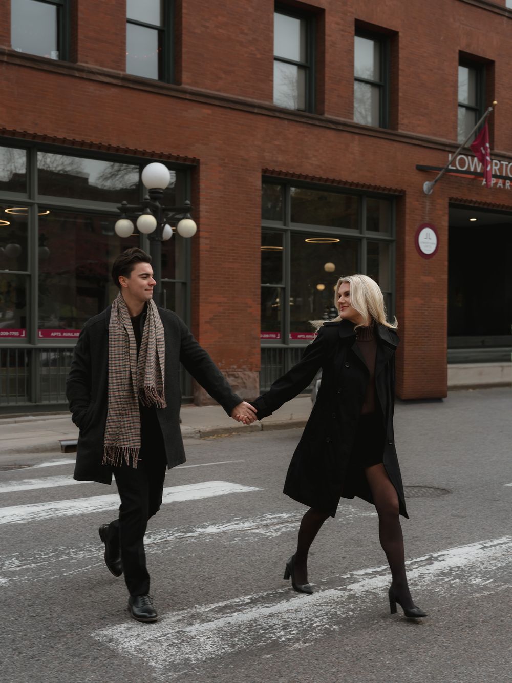A couple holding hands playfully crosses a snowy street in front of a brick building with white lamp posts.