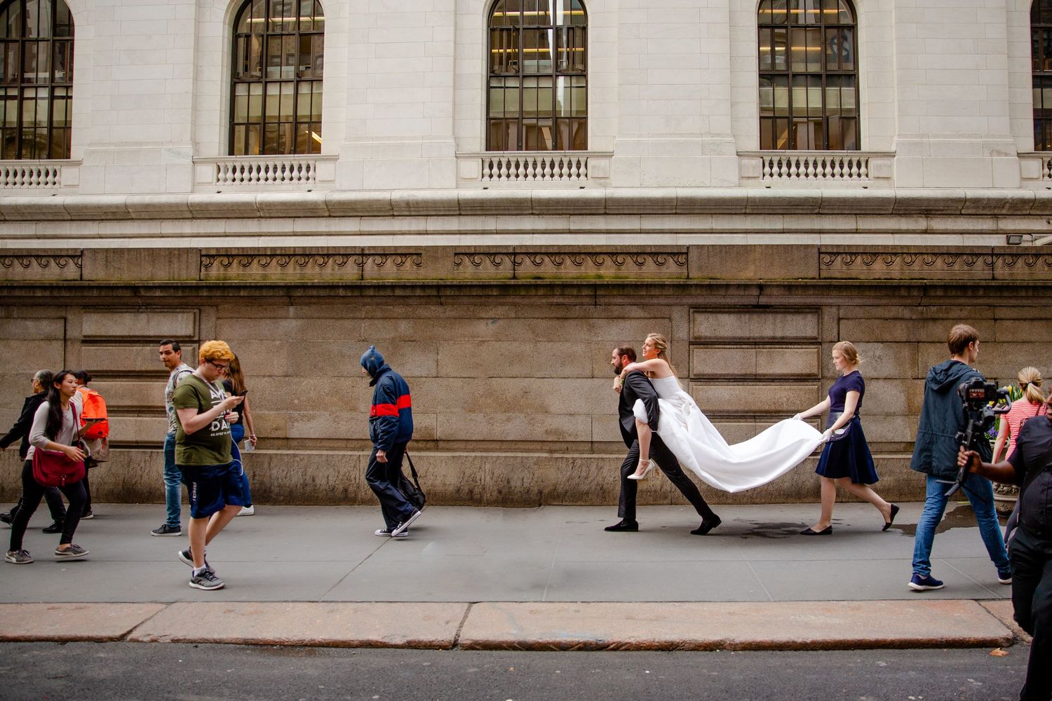 People walking past an ornate stone building with tall windows and columns along the sidewalk.