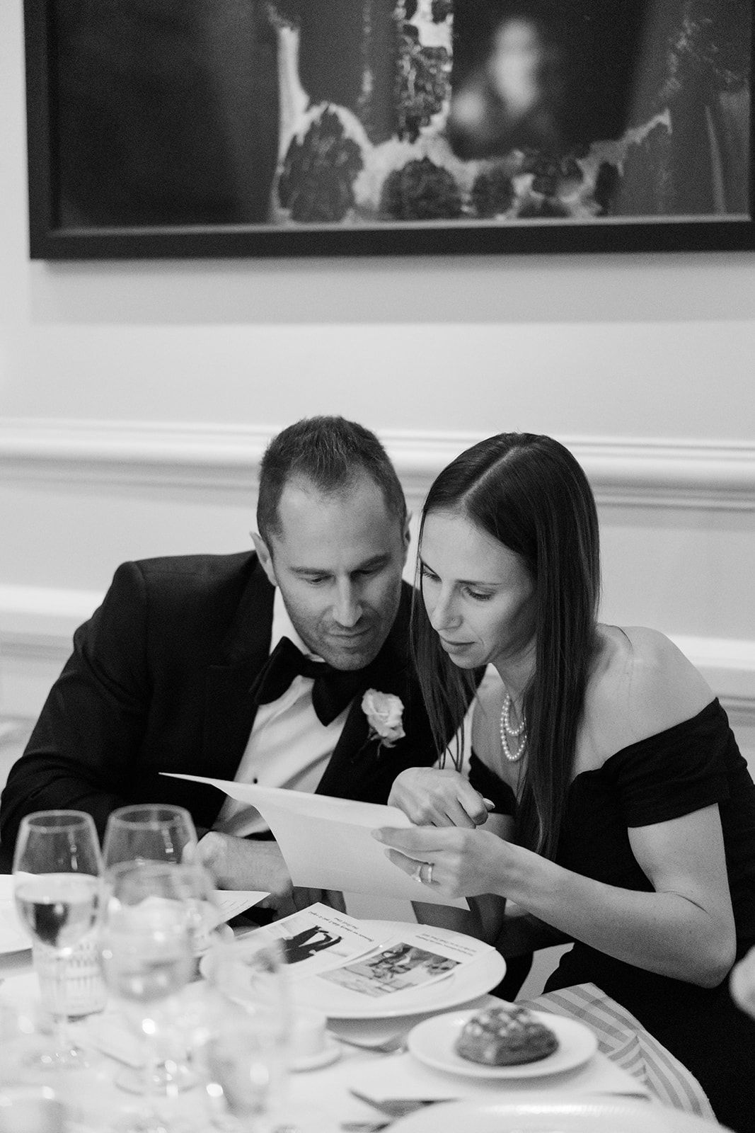 Two people in formal attire lean in close while dining at an elegant restaurant table with white tablecloths and wine glasses.