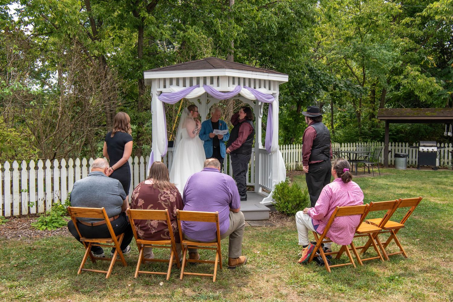 Guests seated in wooden chairs at an outdoor garden wedding ceremony with a white gazebo in the background.