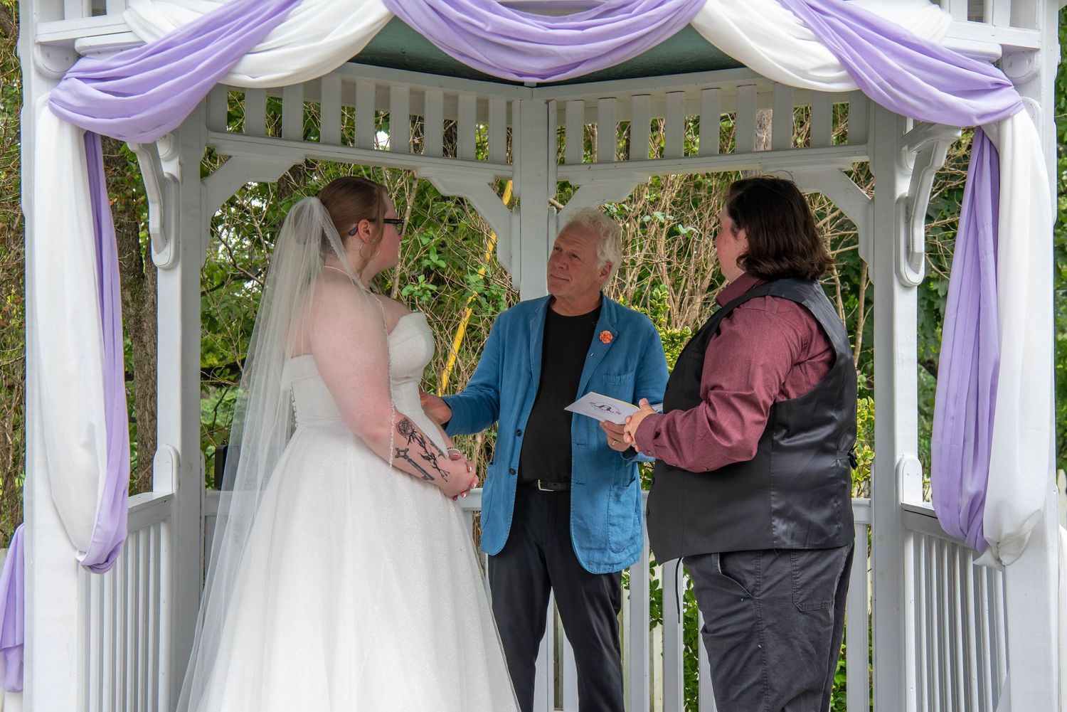 Wedding ceremony taking place under a white gazebo decorated with purple and white fabric draping.