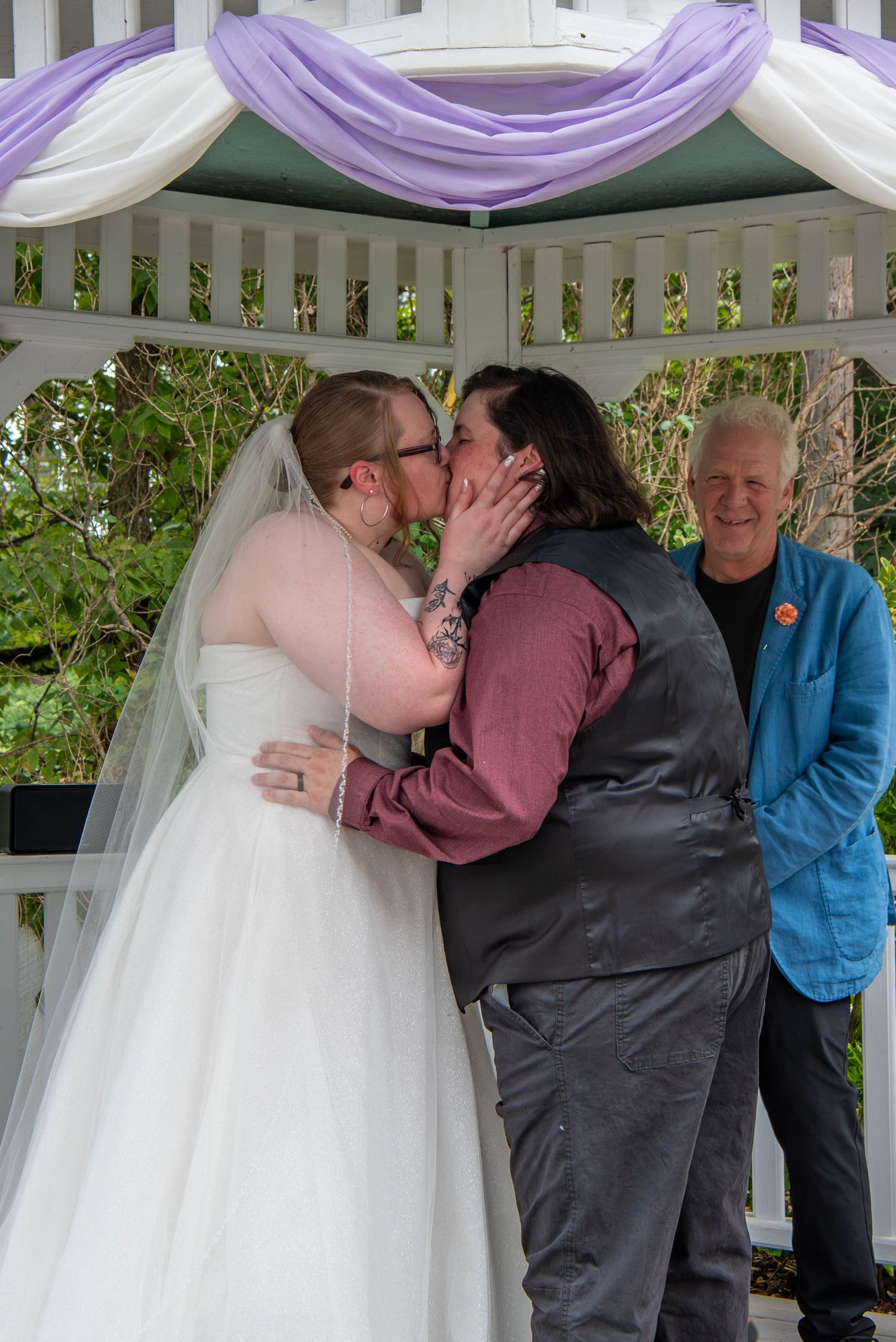 A wedding ceremony takes place under a white gazebo decorated with purple fabric draping.