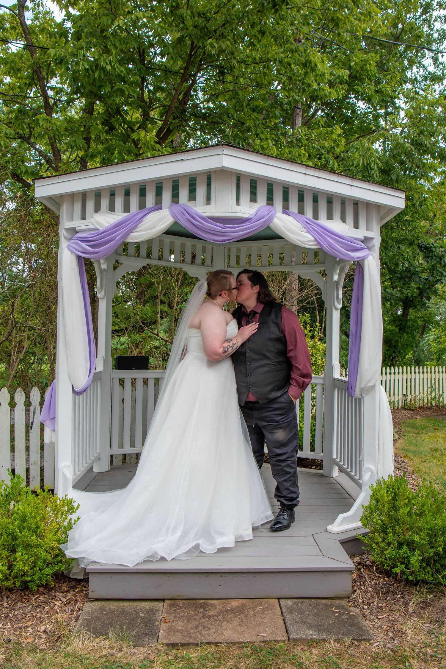 A romantic wedding scene in a white gazebo decorated with purple fabric draping and surrounded by green trees.