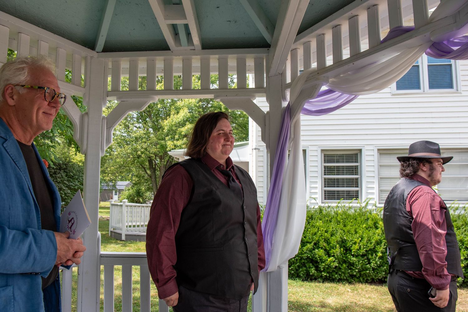 Three people stand under a white gazebo on a sunny day with green landscaping visible in the background.