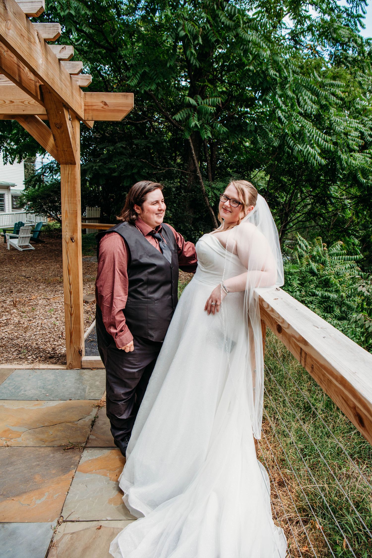 A wedding couple shares a candid moment under a wooden pergola surrounded by greenery.