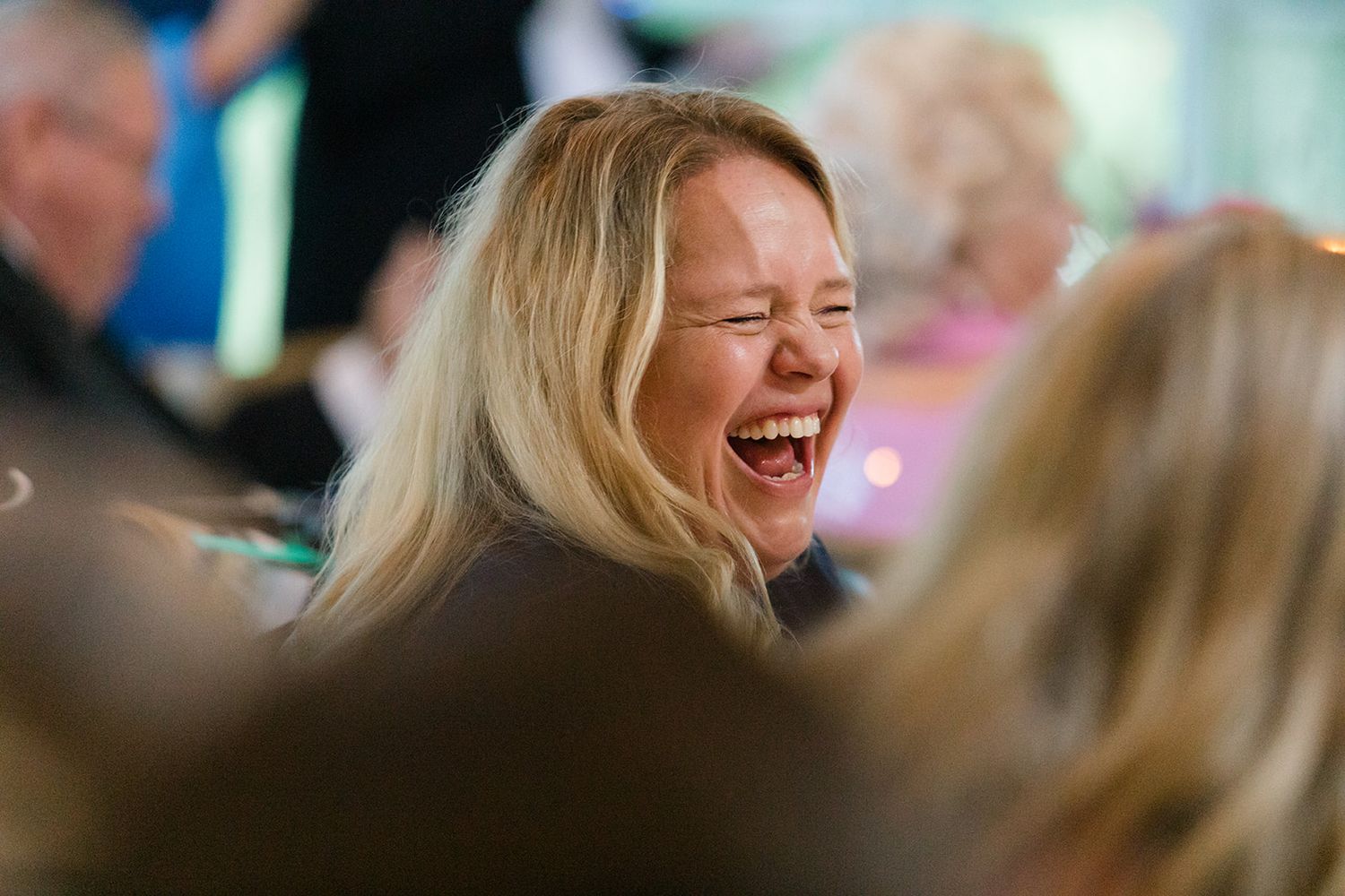 A person with blonde hair laughs joyfully in a candid moment during a social gathering.