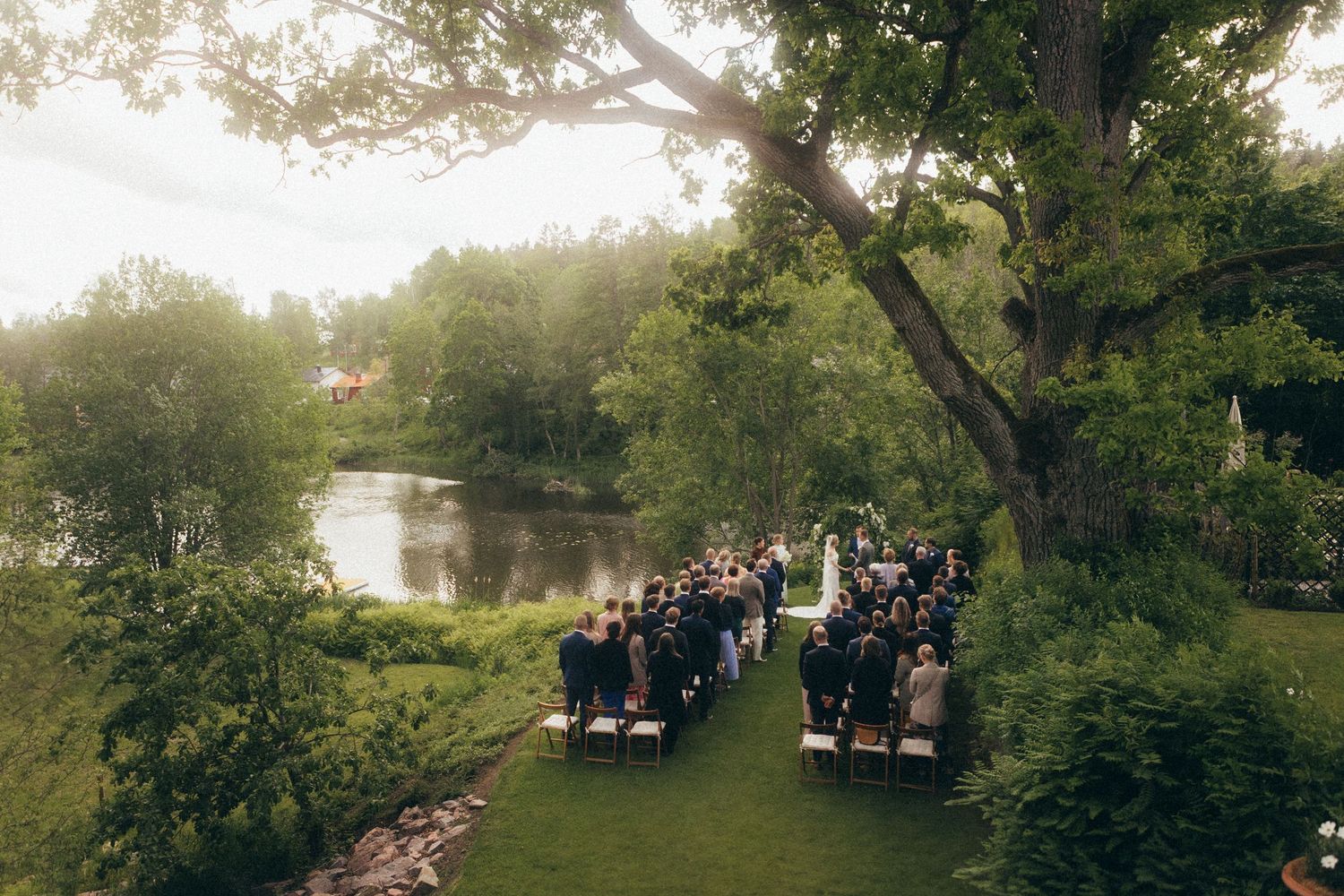 Outdoor wedding ceremony setup on a lush green lawn beside a tranquil pond with guests seated in white chairs.